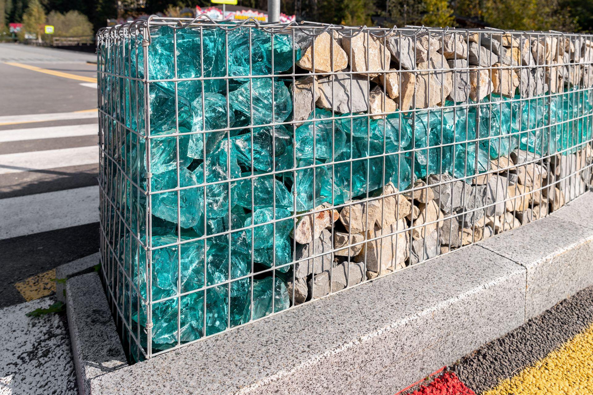 Detailaufnahme eines neuen, modernen Gabionenzauns mit Metallkäfig, der mit Schotter und zerbrochenen blauen Kunstglassteinen gefüllt ist. Stadt Straße Straße Bürgersteig Schutz